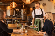 Die Heimat schmecken kann man bei den Allgäuer Genusstagen vom 19. September bis 4. Oktober. Foto: Landkreise Ost- und Unterallgäu / Studio Zuchtriegel Auf dem Foto sitzen zwei Frauen am Tisch. Eine Kellnerin im Dirndl reicht gerade das Essen.