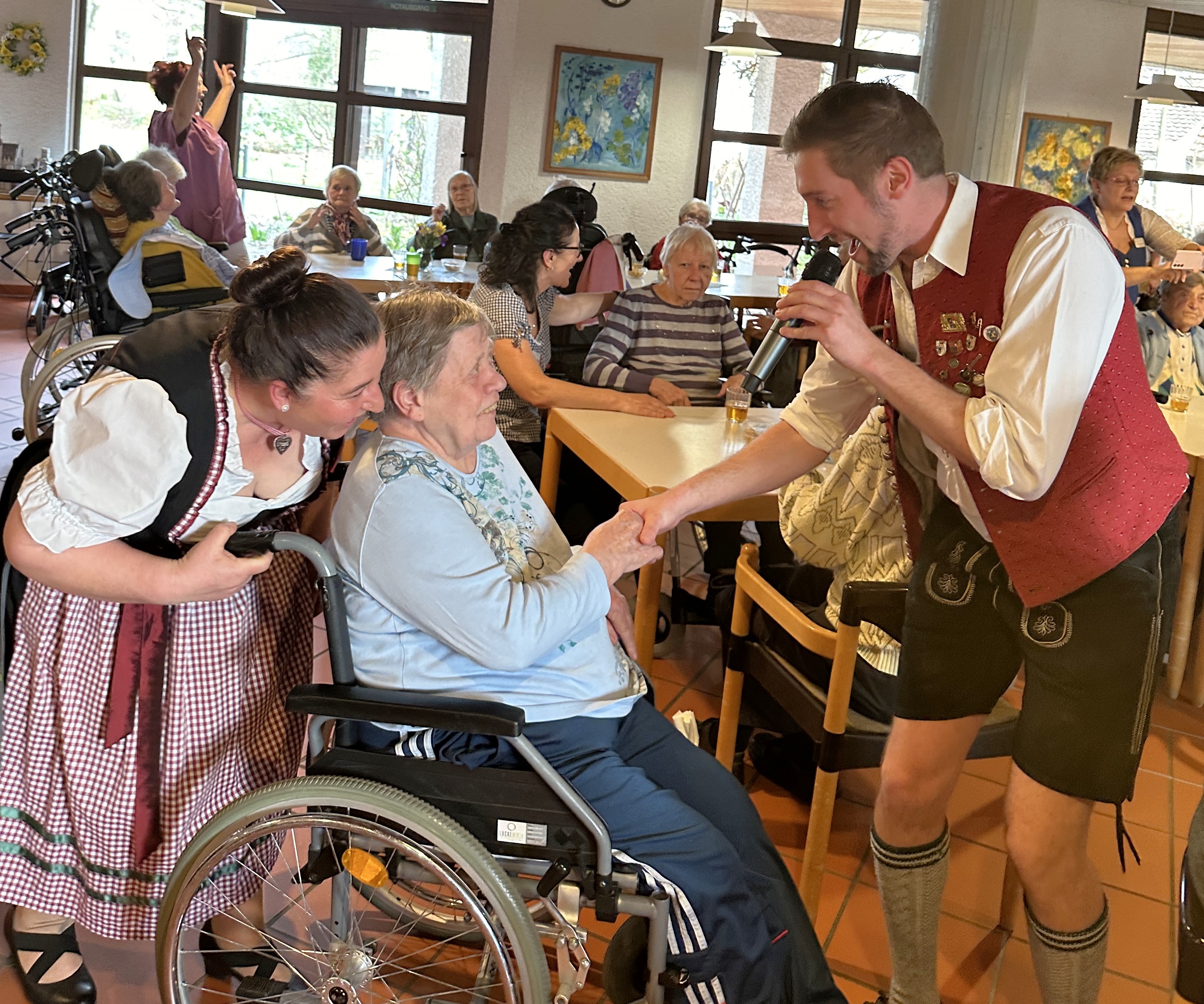Eine Seniorin im Rollstuhl reicht dem Sänger der Musikkapelle die Hand. Hinter der Dame steht eine Frau im Dirndl. 